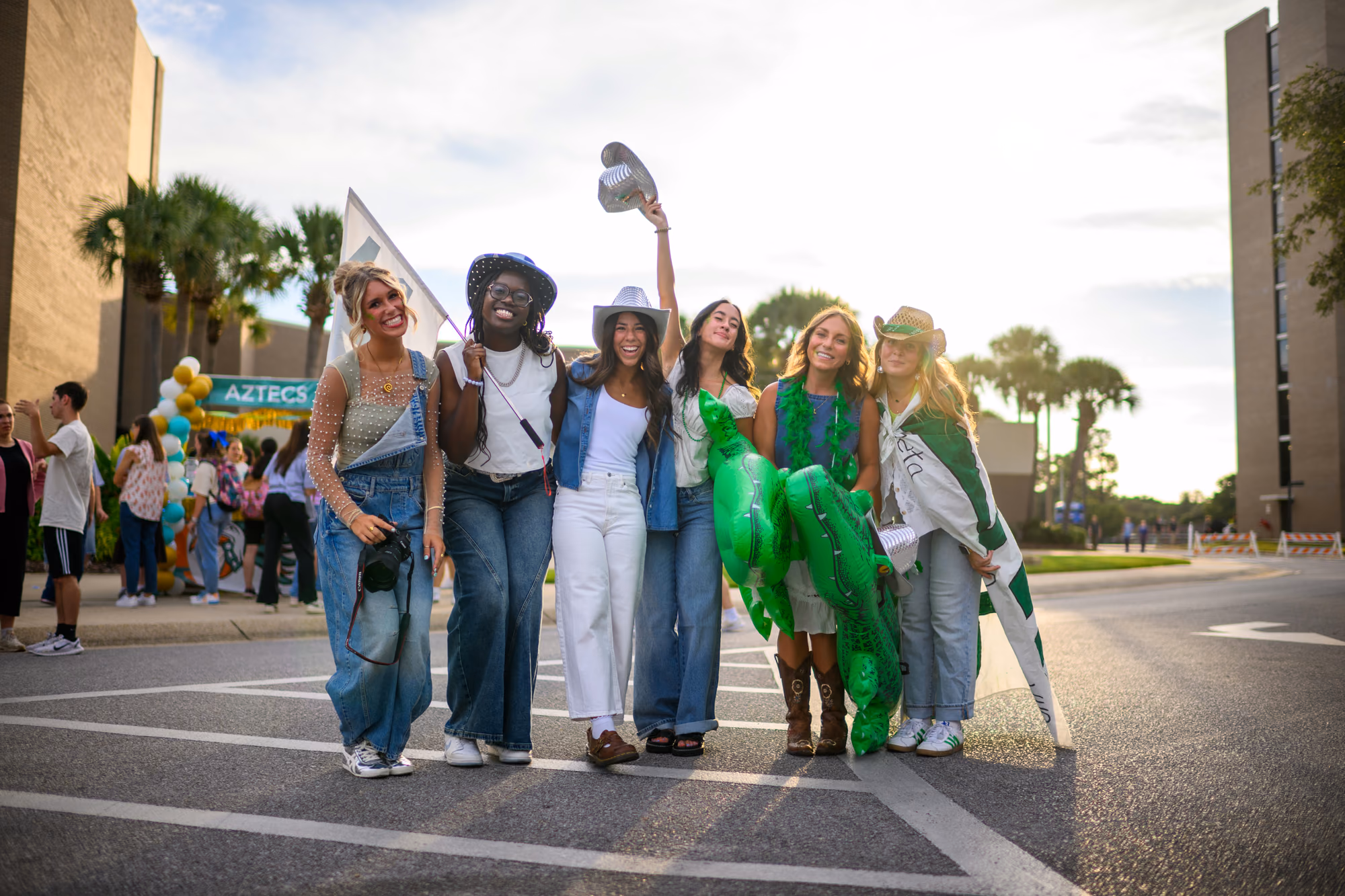 Six friends in colorful costumes pose on a campus street, one holding a green inflatable cactus