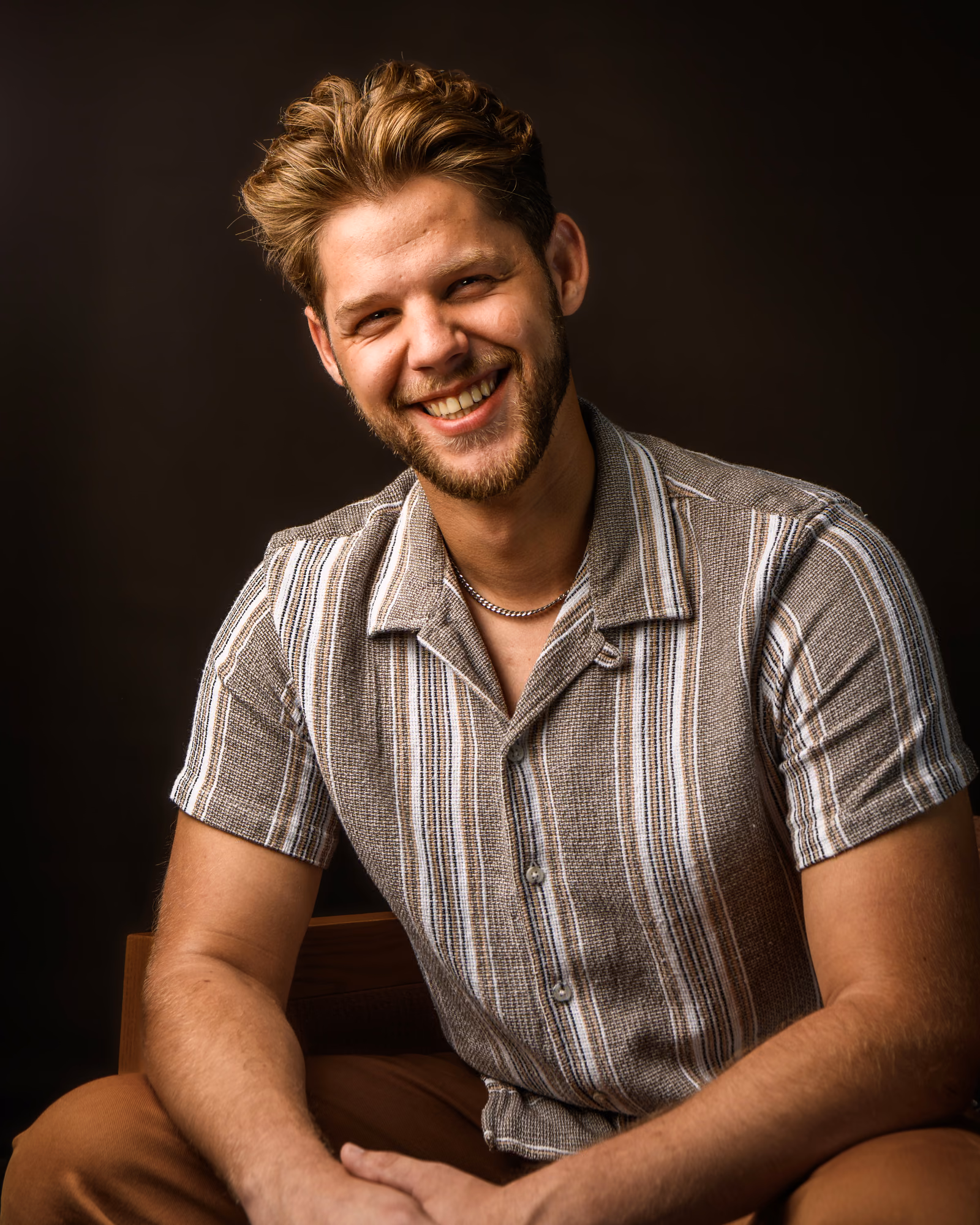 A smiling man in a striped shirt seated against a warm, dark studio backdrop
