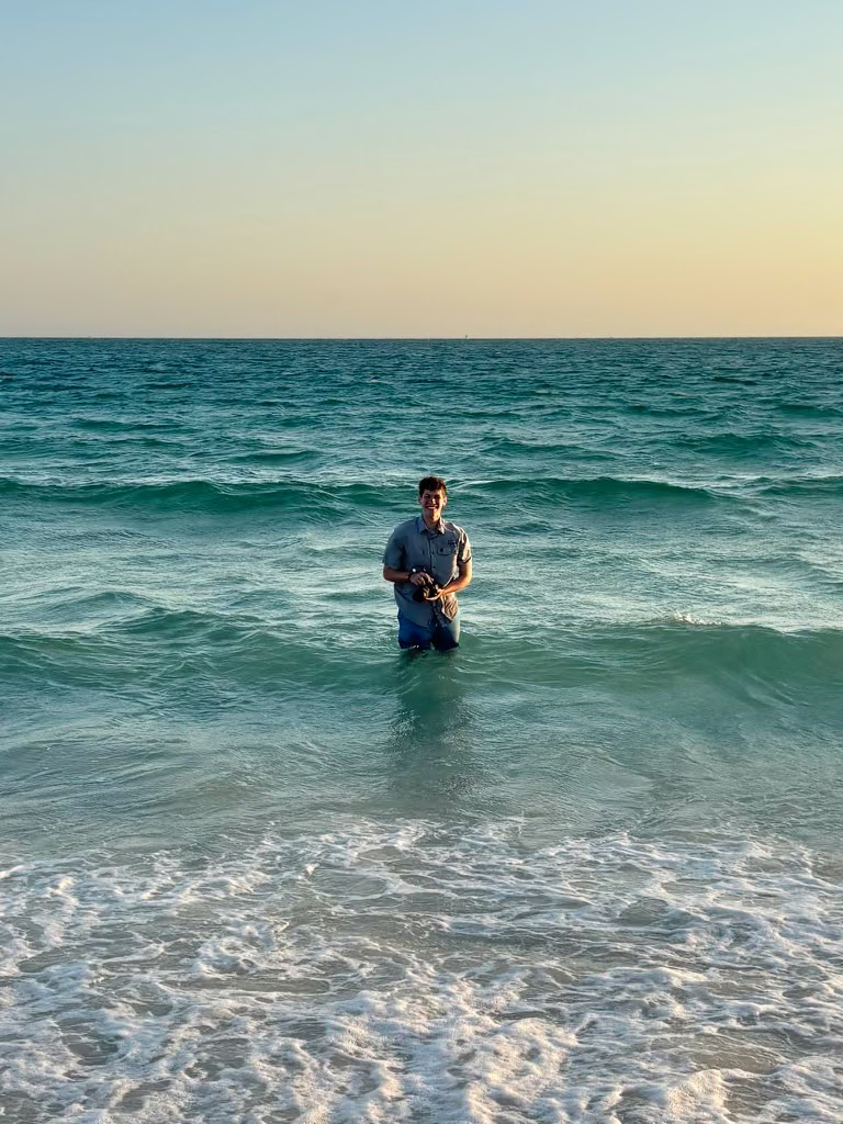 William Lund holding a camera while standing in ocean surf beneath a pale evening sky