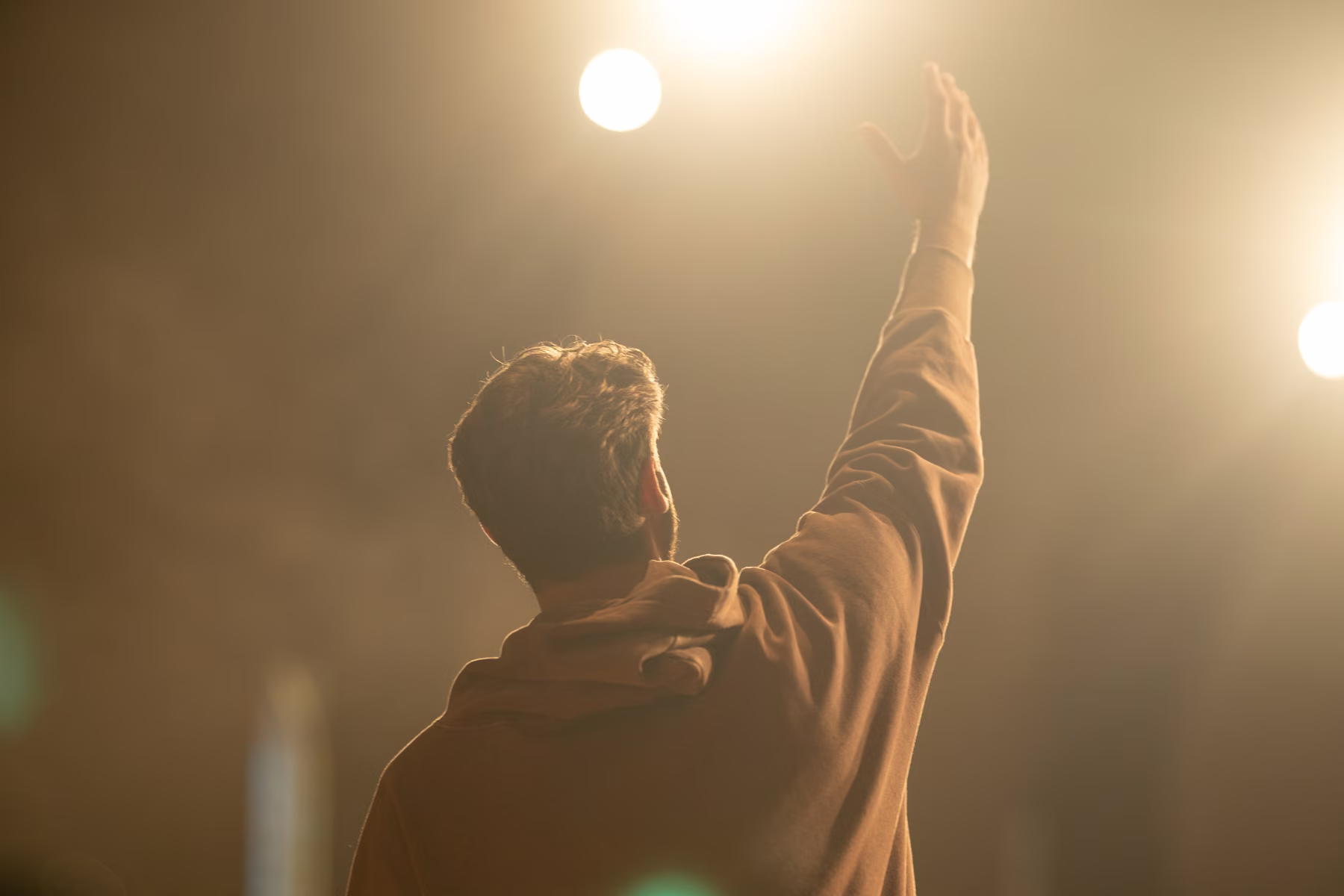 Alex seen from behind with one hand raised in golden light and haze.