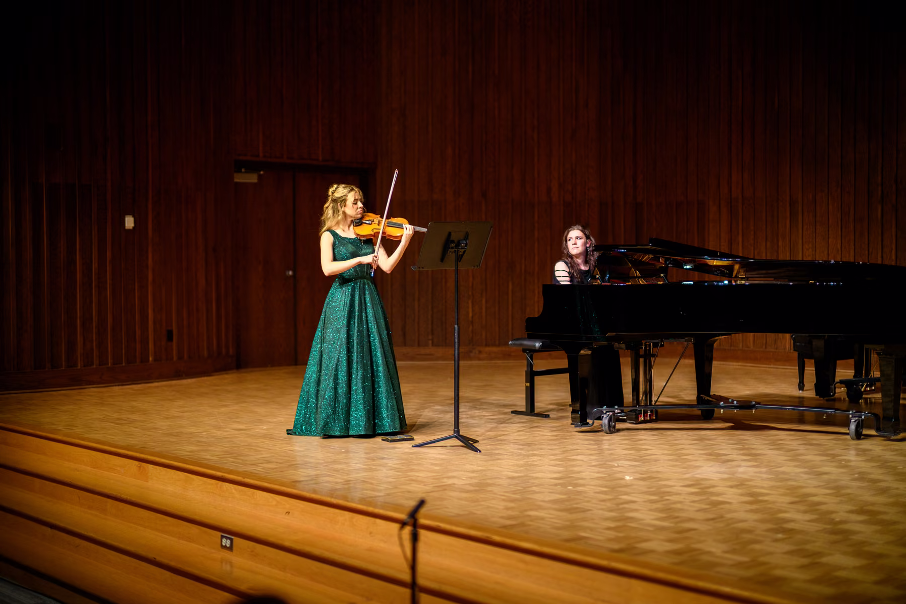 Alyssa playing violin on a wooden stage beside a grand piano.