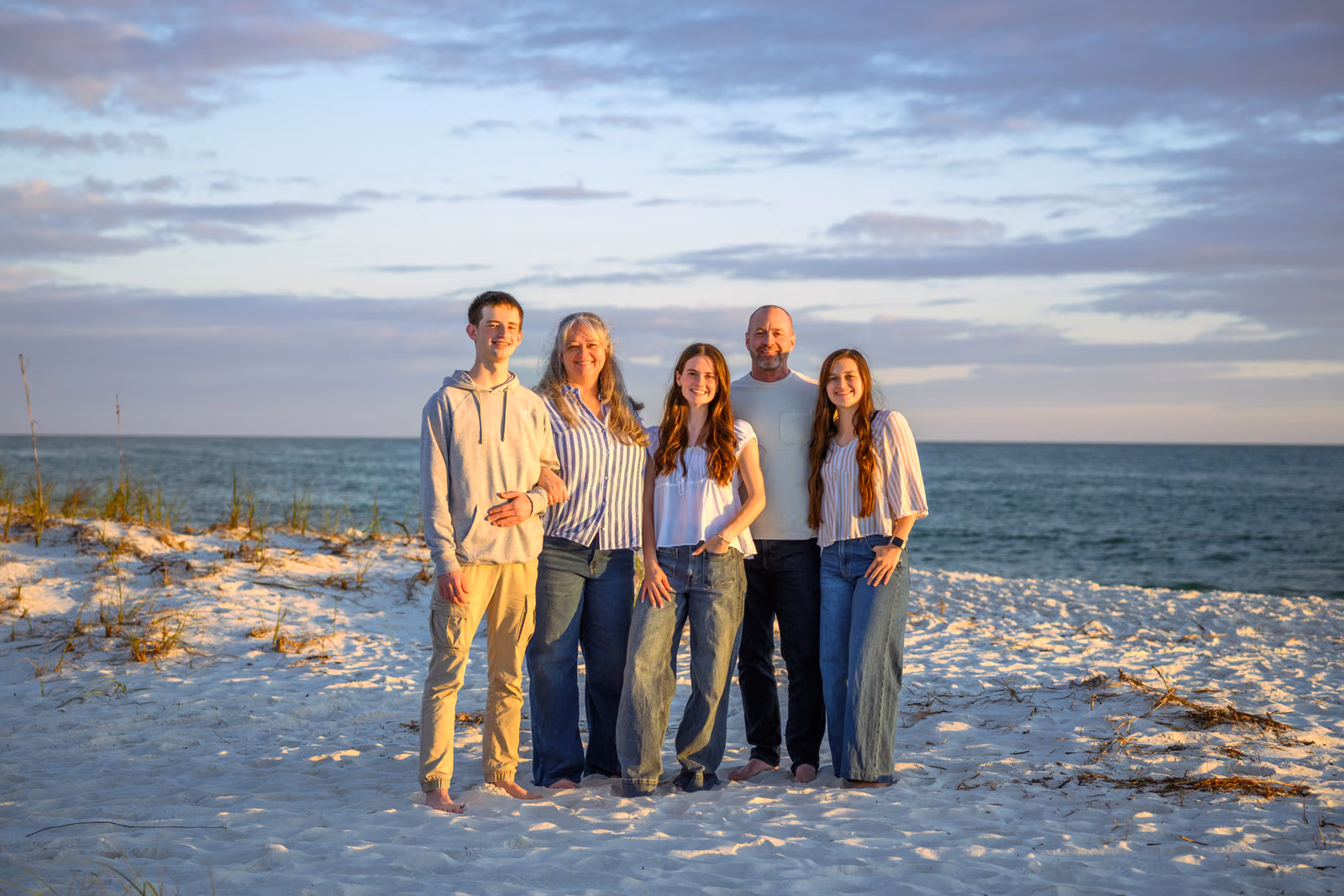 A family of five smiles for a portrait on pale sand with the ocean behind them.