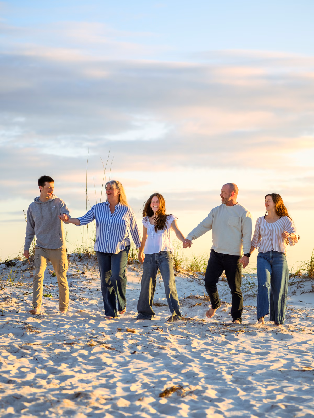 Five family members hold hands and walk across the sand together beneath a pastel evening sky.