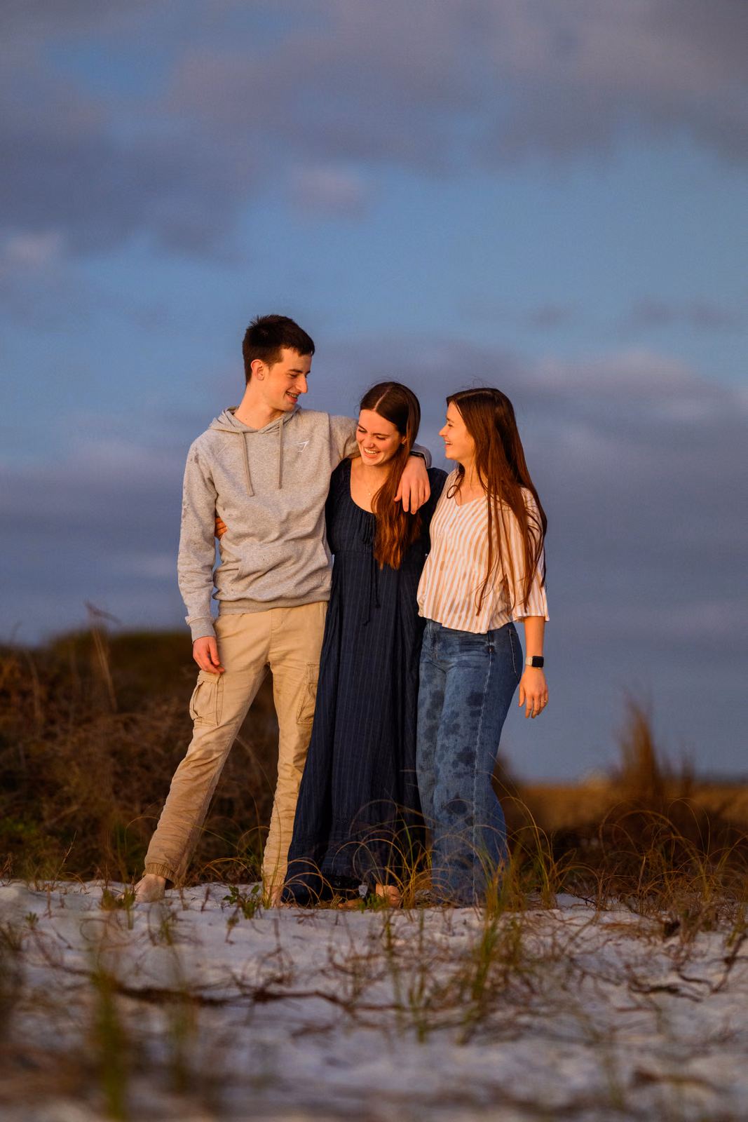 Three people standing close together and smiling among sandy dunes beneath a blue evening sky.