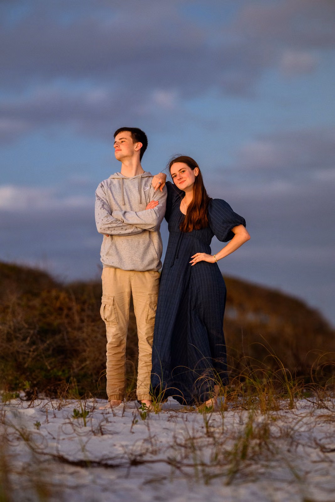 Bailey in a navy dress poses beside a family member in a gray hoodie among the evening dunes.
