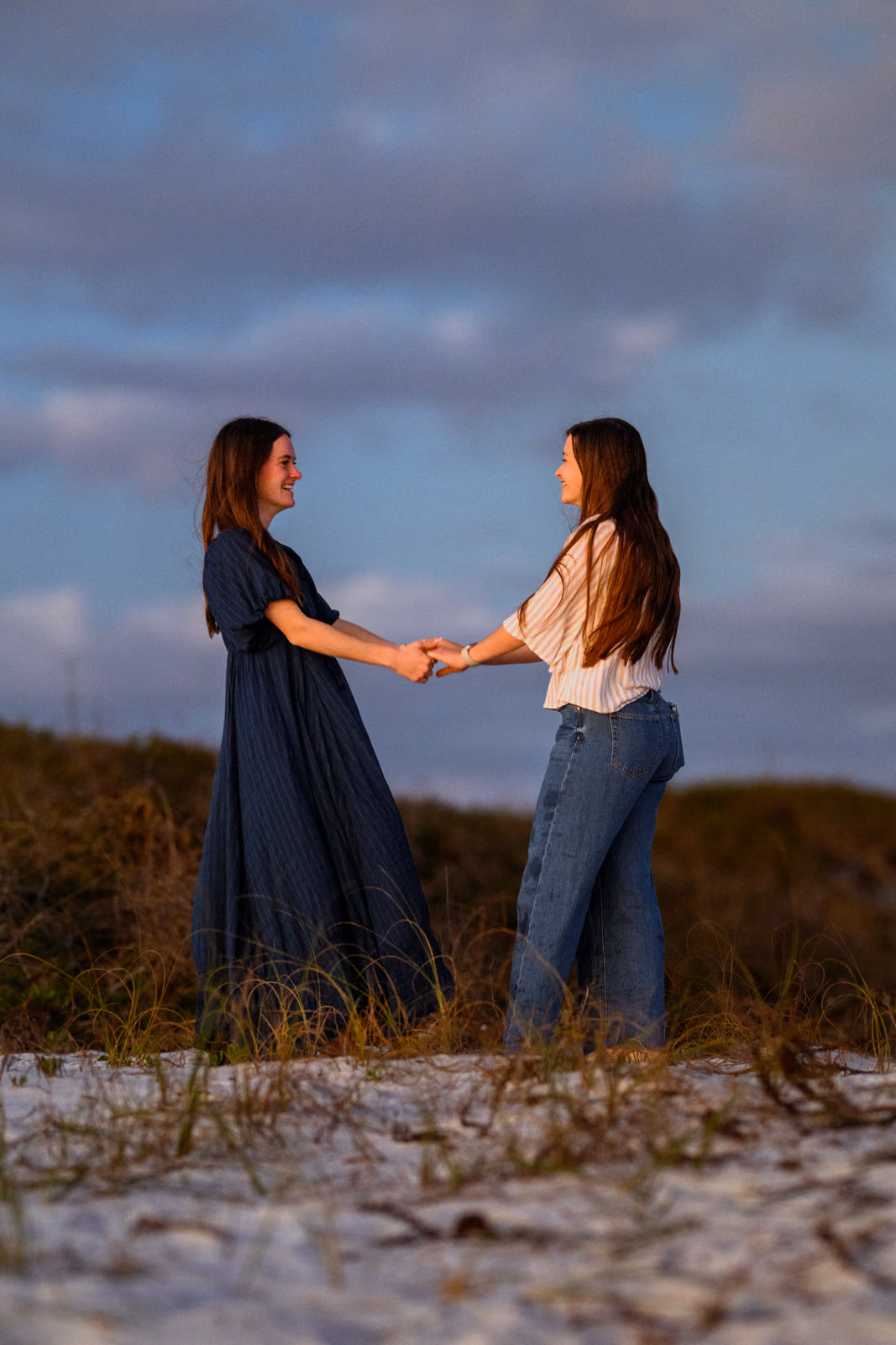 Two people holding hands and facing each other among sandy dunes beneath a blue evening sky.