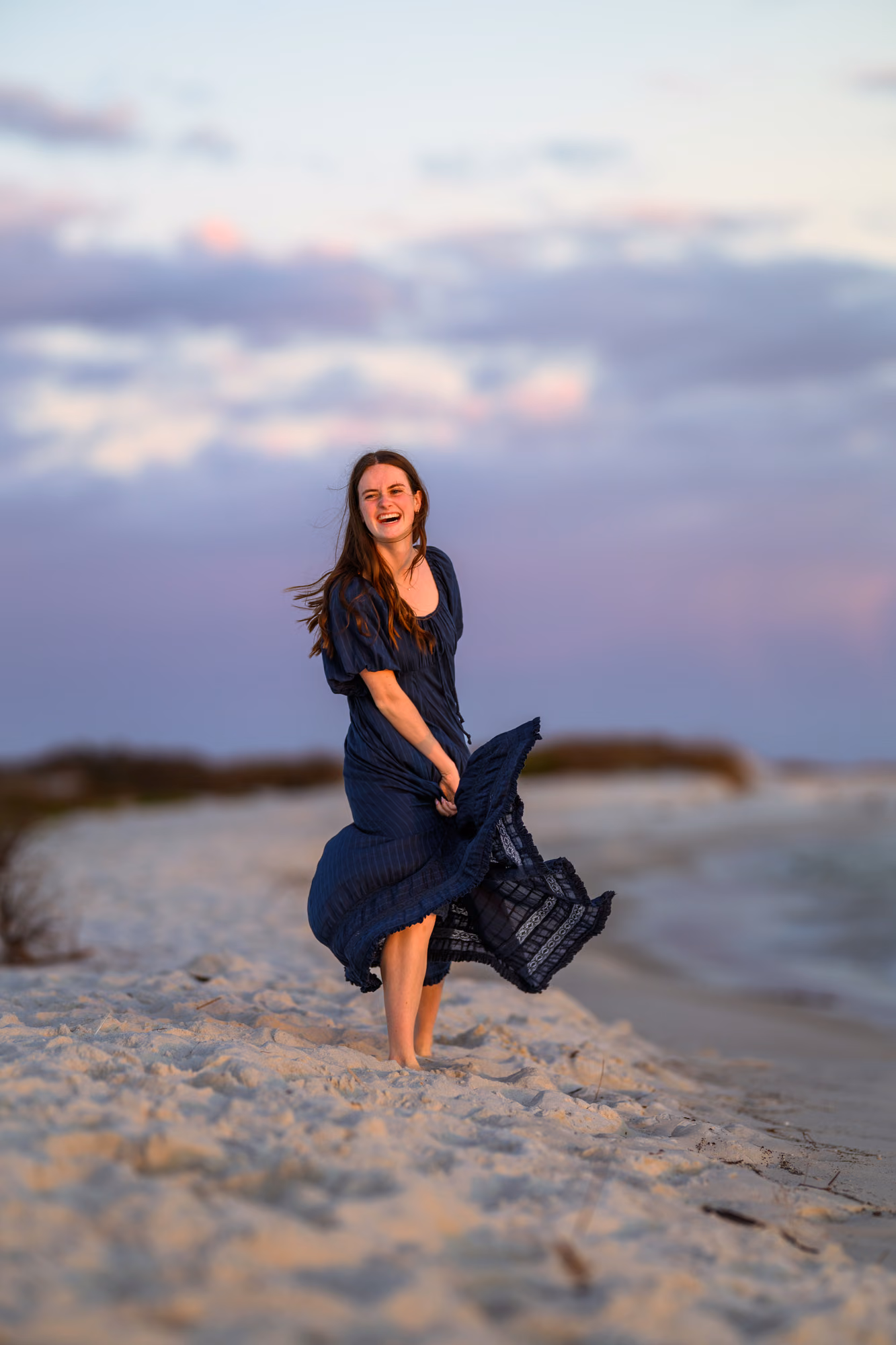 Bailey in a navy dress smiling and lifting the hem while standing barefoot on a beach.