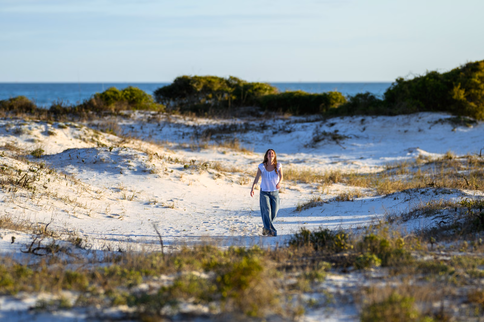 Bailey in a white blouse and jeans walks through a wide landscape of pale dunes.