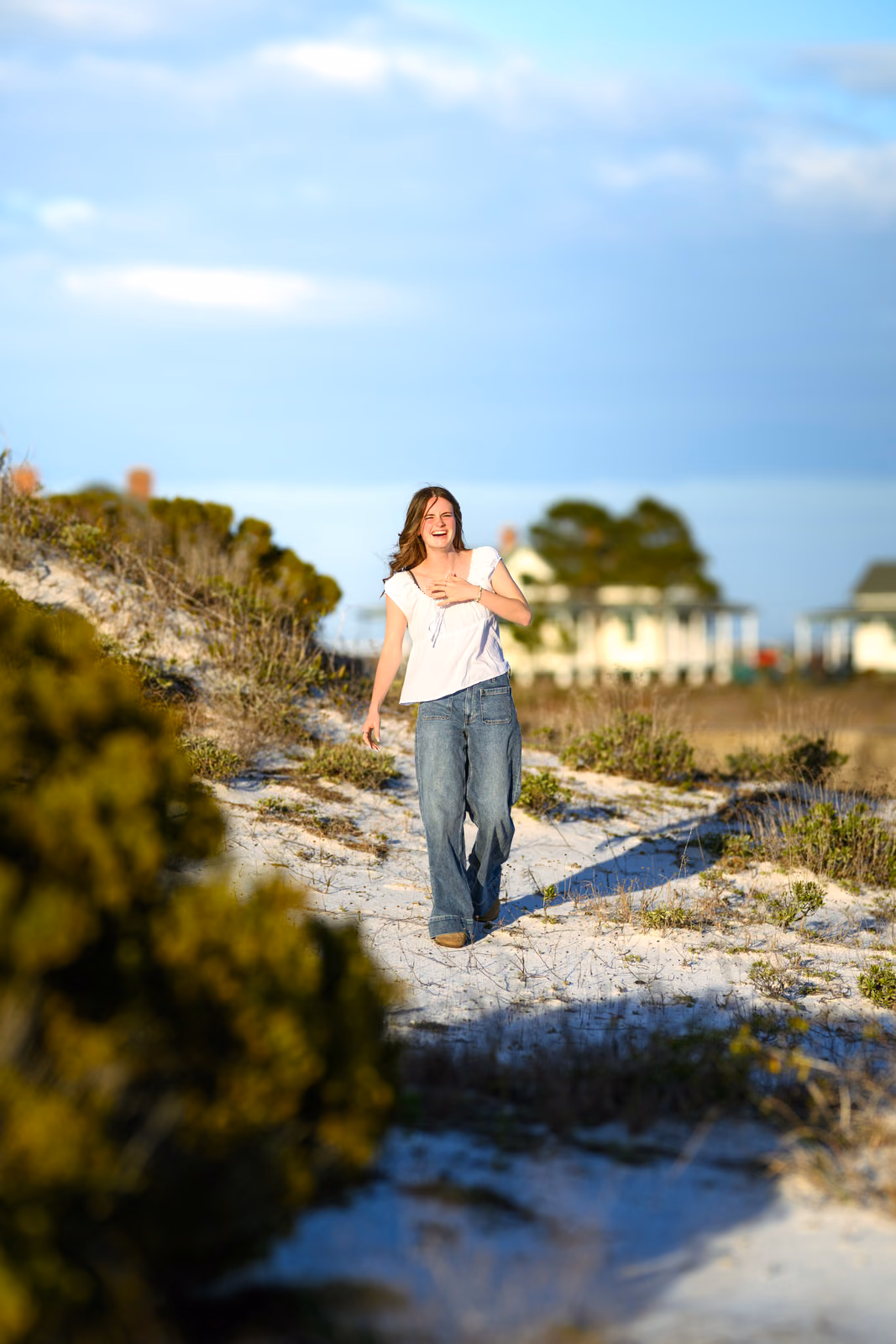 Bailey laughs while walking through sunlit dunes in a white blouse and jeans.