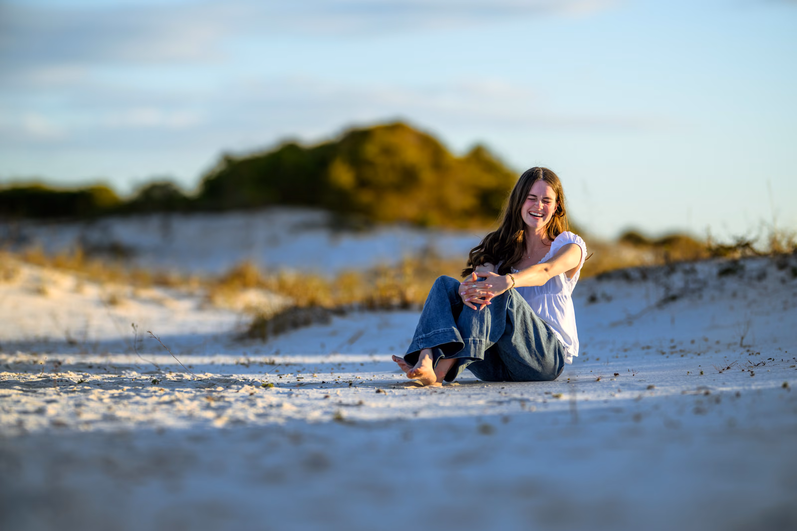 Bailey in a white blouse and jeans sits laughing on the sand.