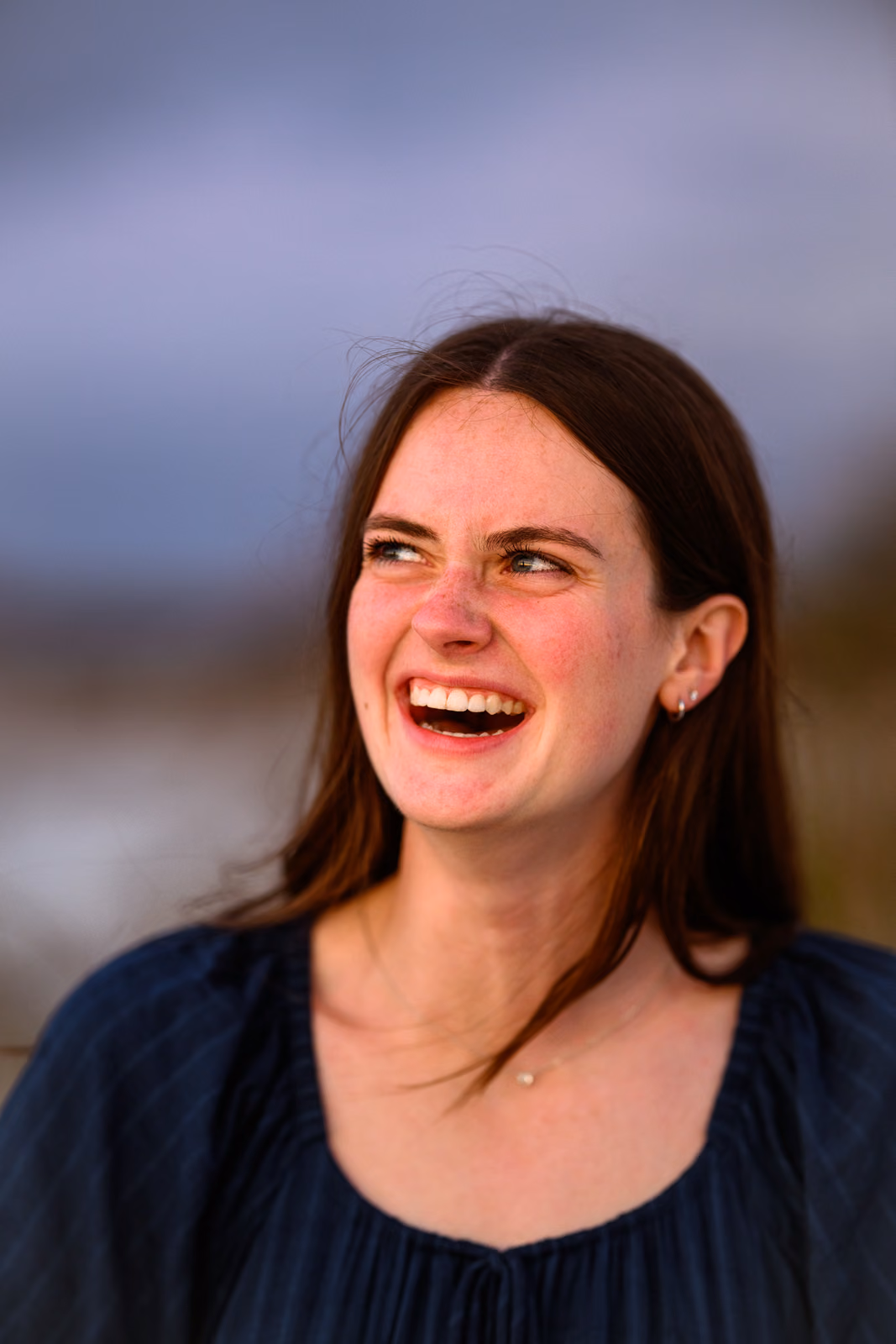 Bailey in a navy dress laughing and looking upward against a softly blurred beach background.
