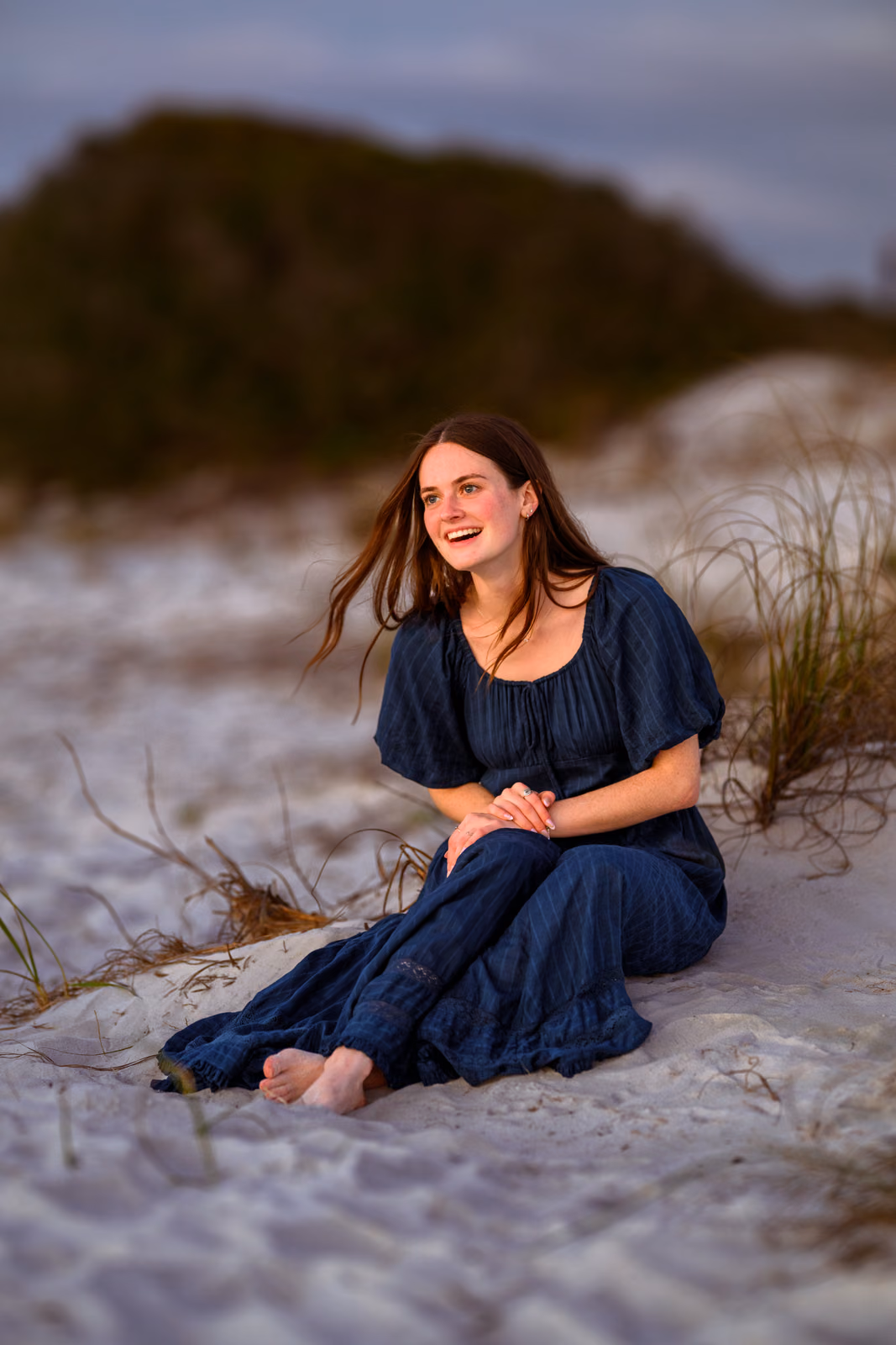 Bailey in a navy dress sitting barefoot in pale sand beside dune grass and smiling to one side.