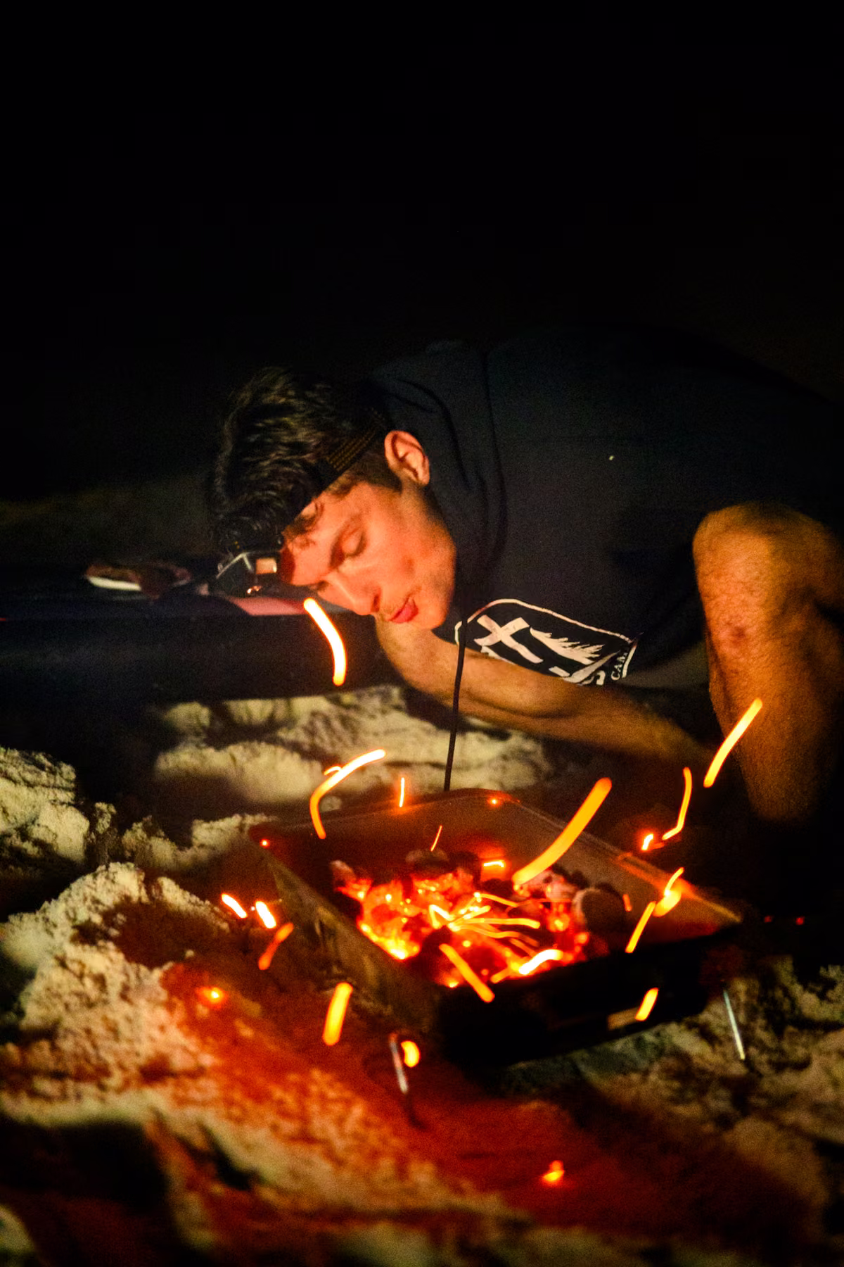 Noah blows on glowing charcoal as orange sparks rise from a small grill on the sand.