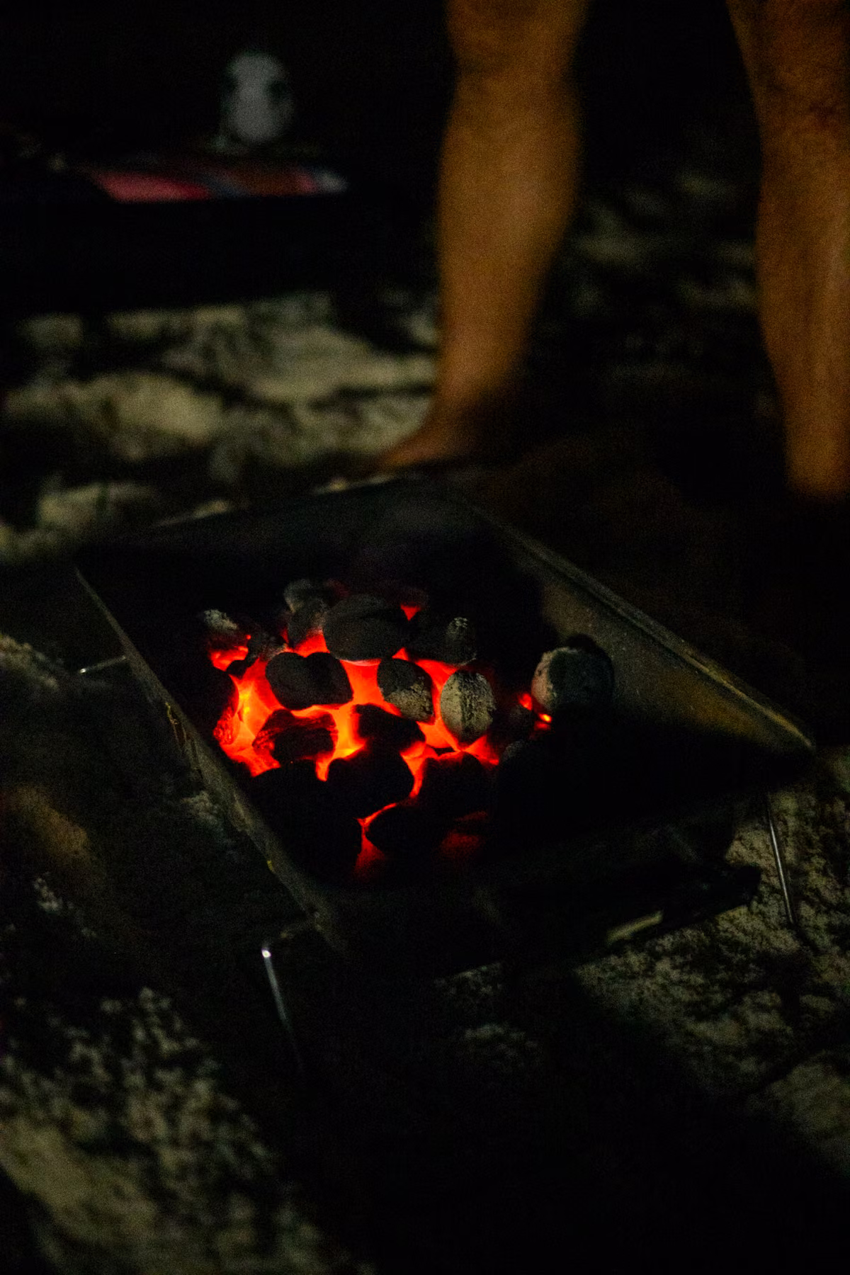 Red-hot charcoal glows inside a small grill resting on the sand at night.