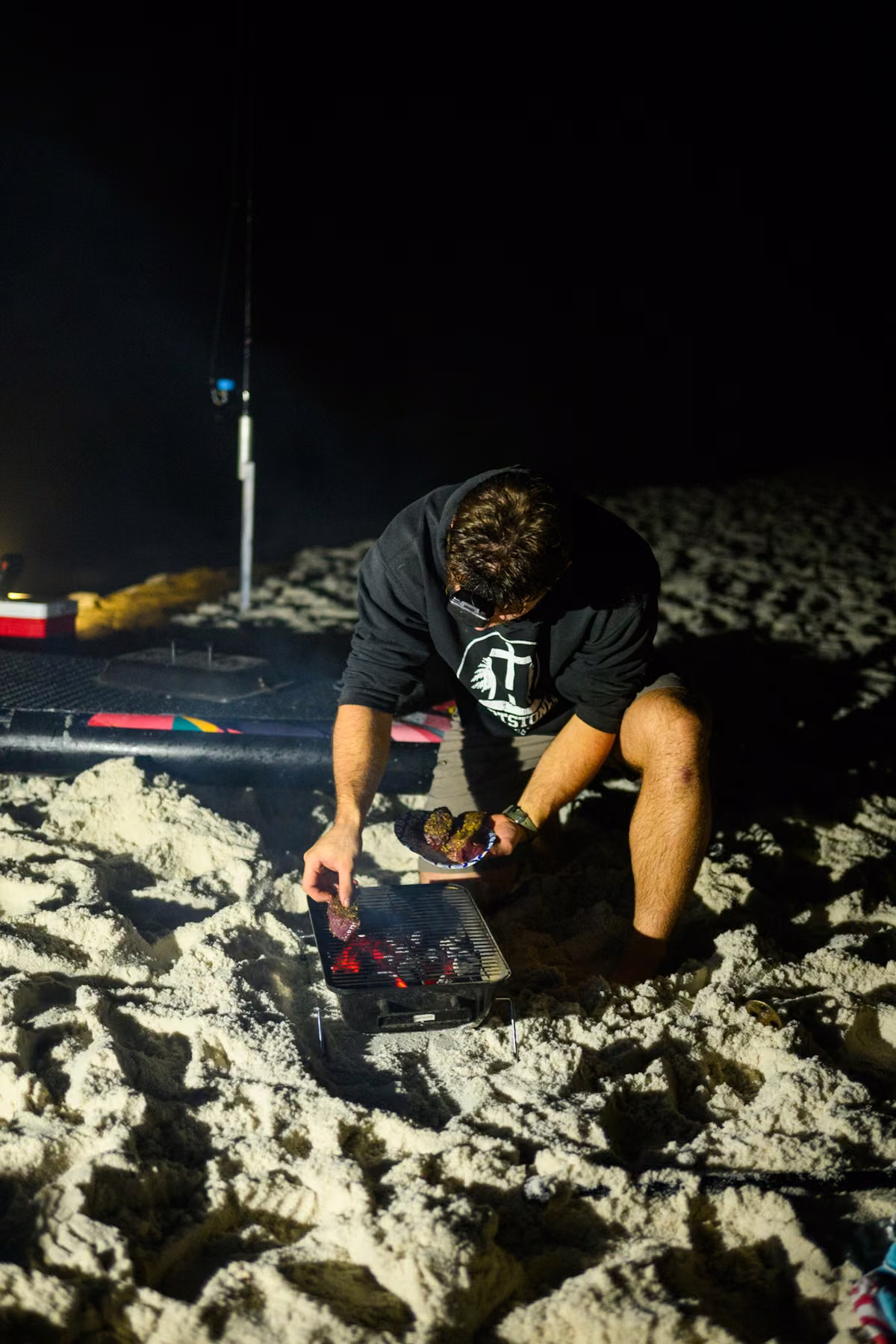 Noah kneels on the sand and places seasoned steaks on a small charcoal grill.