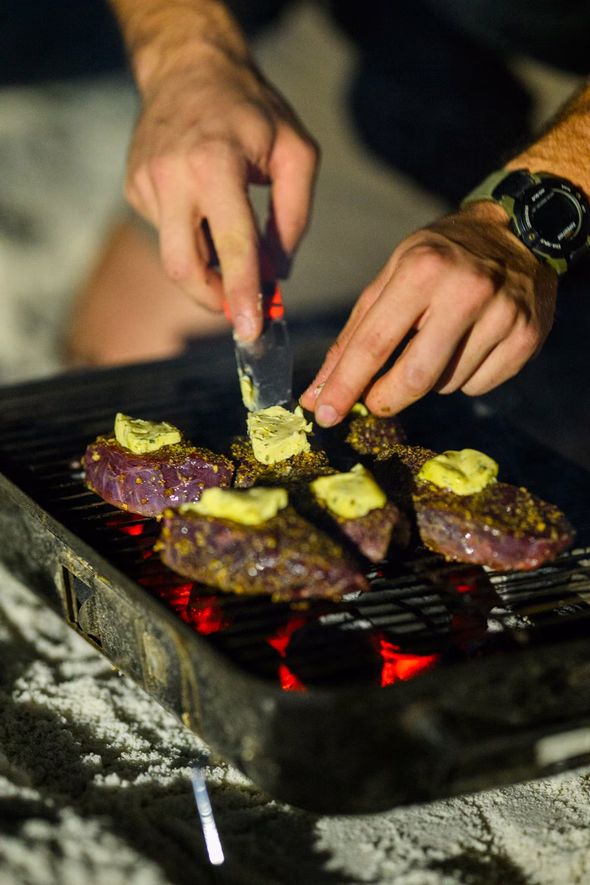 Close-up of Noah adding herb butter to steaks over glowing coals.