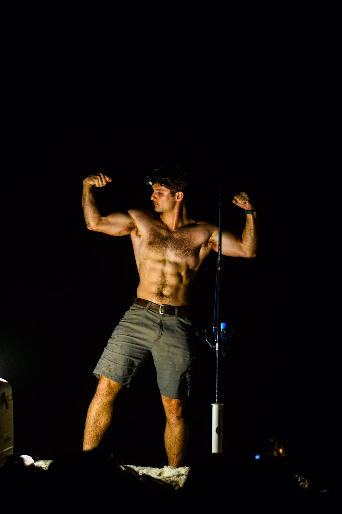 Noah playfully flexes beside an upright fishing rod while wearing a headlamp on the beach at night.