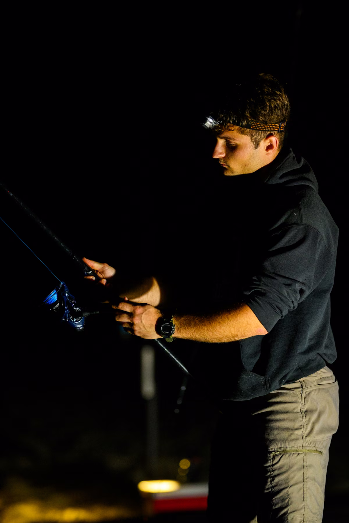 Noah adjusts a blue fishing reel while wearing a headlamp in the darkness.