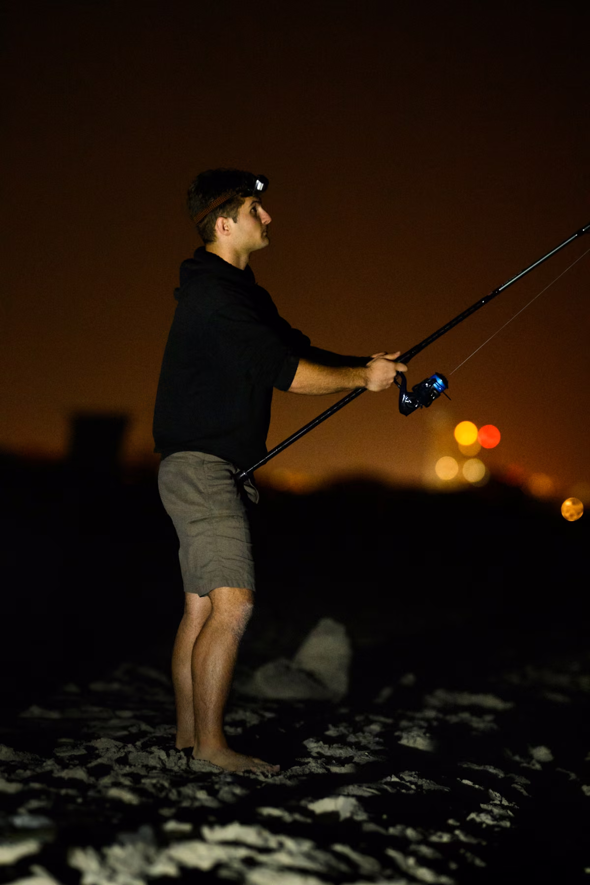 Noah stands barefoot on the sand holding a fishing rod against distant amber lights.