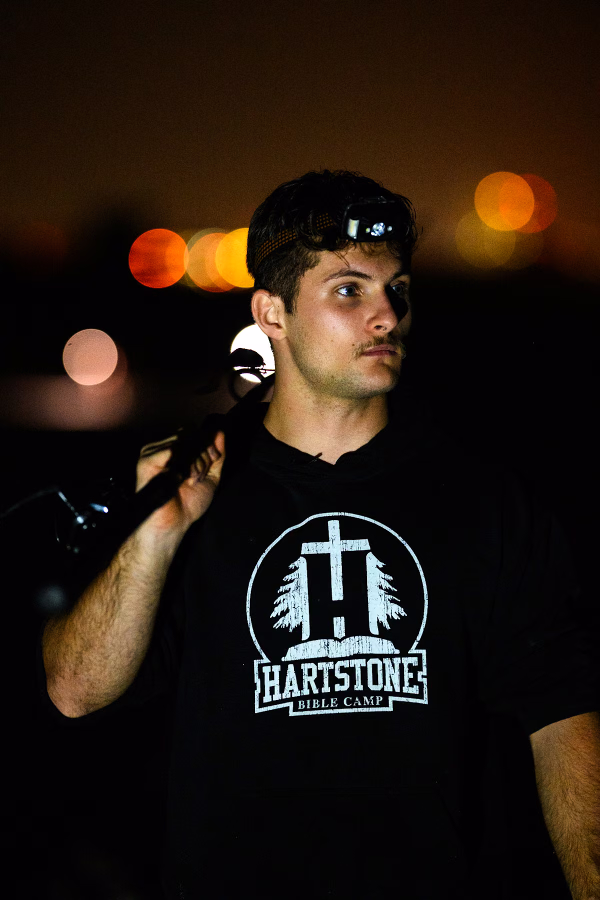 Noah rests a fishing rod on his shoulder beneath a headlamp, with warm lights blurred behind him.