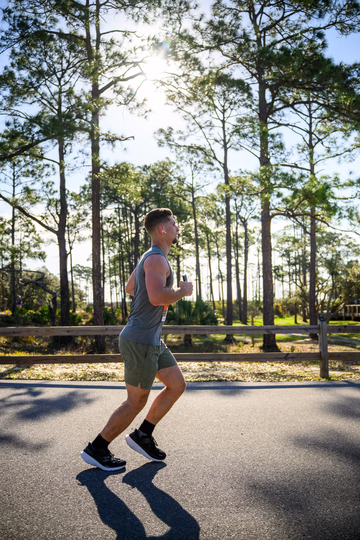 Side view of Noah running past a wooden fence beneath sunlit pine trees.