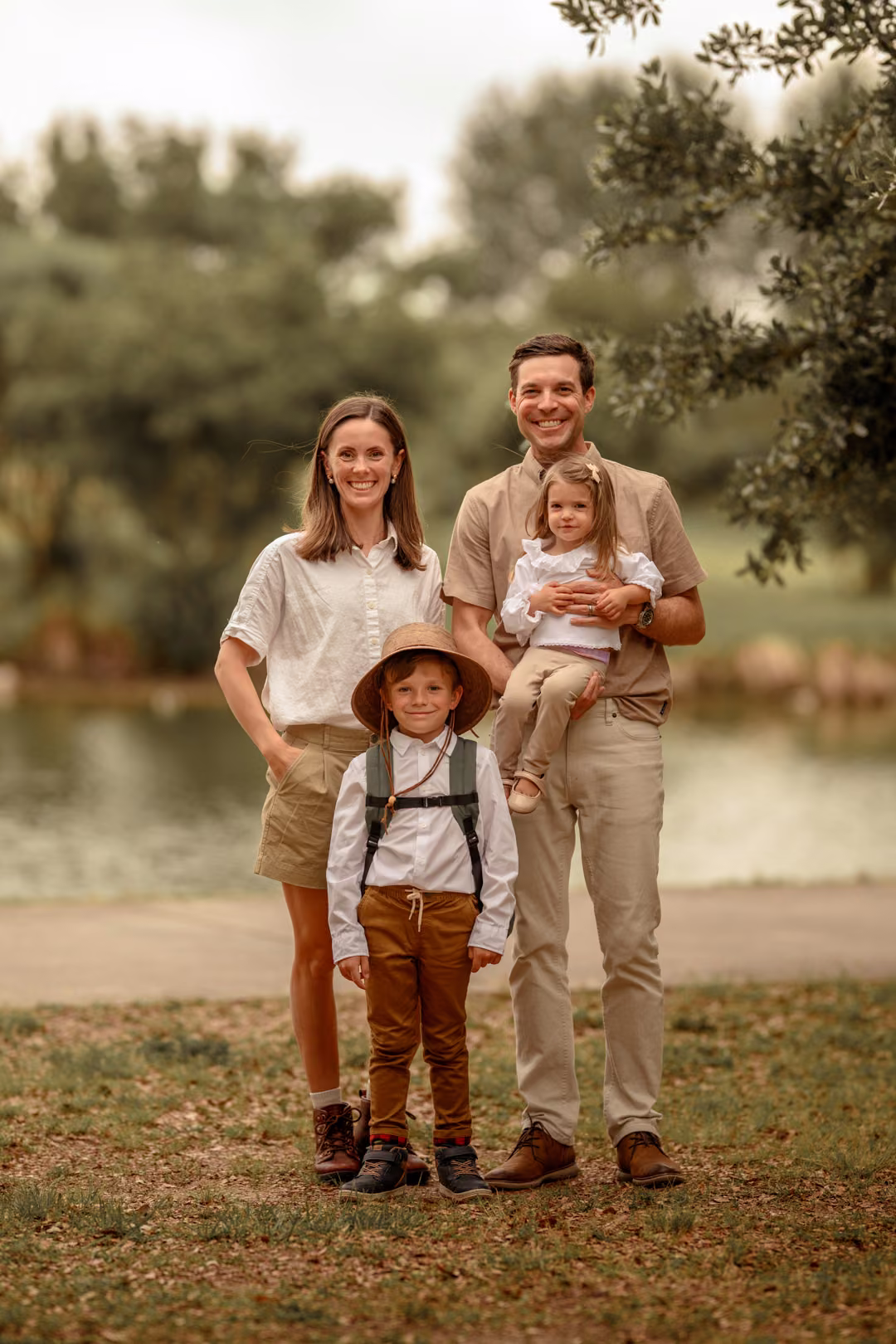 A family of four standing together beside a lake, dressed in coordinated warm neutral colors.