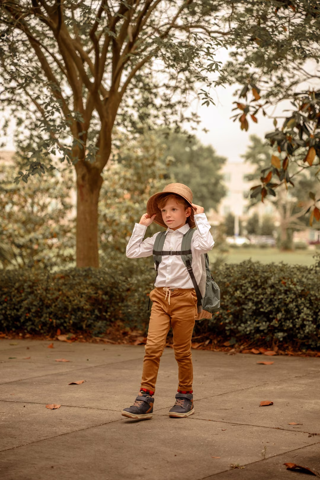 A young boy wearing a backpack and holding the brim of his explorer-style hat beneath a tree.
