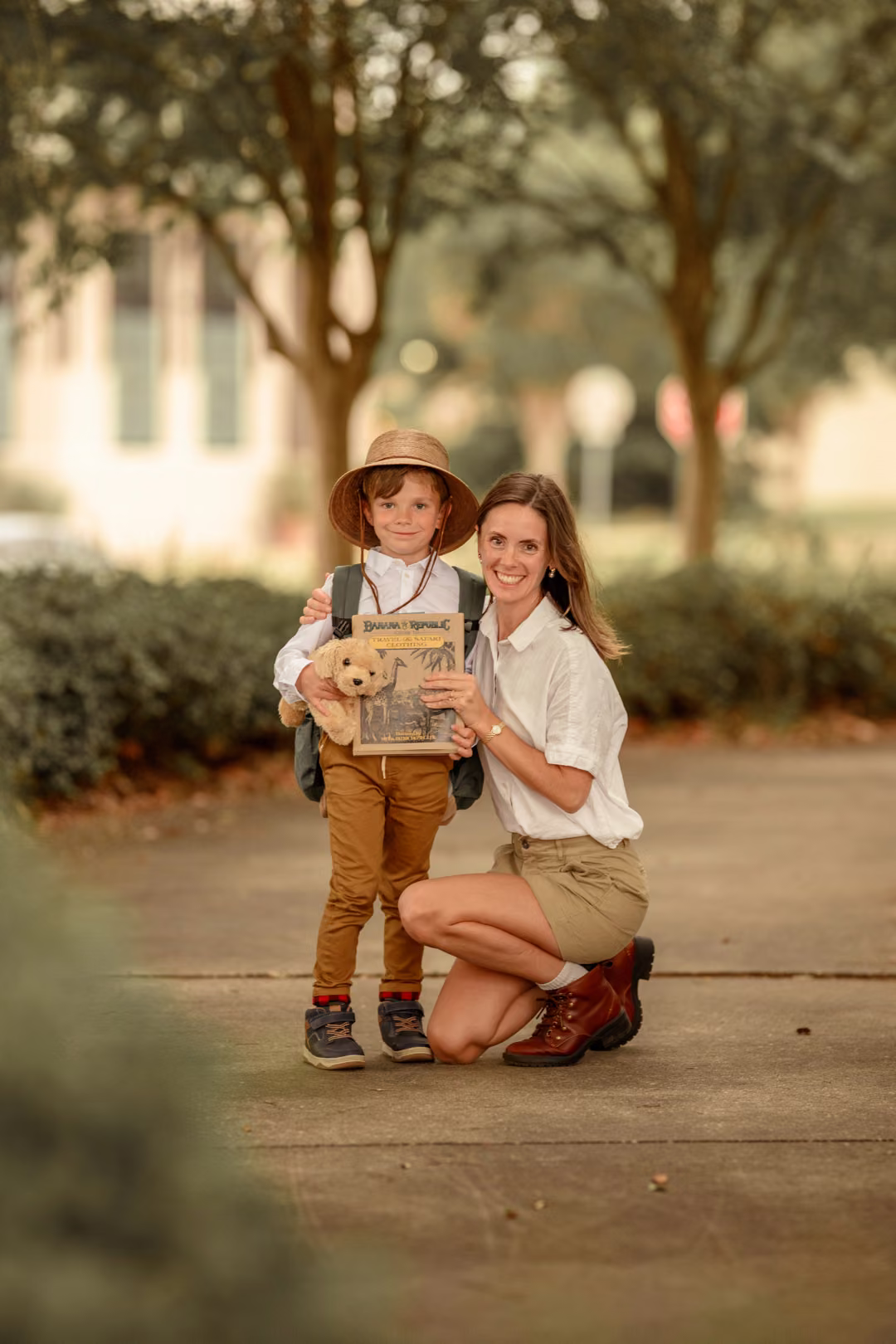 A mother kneeling beside her son as he holds a teddy bear and a book.
