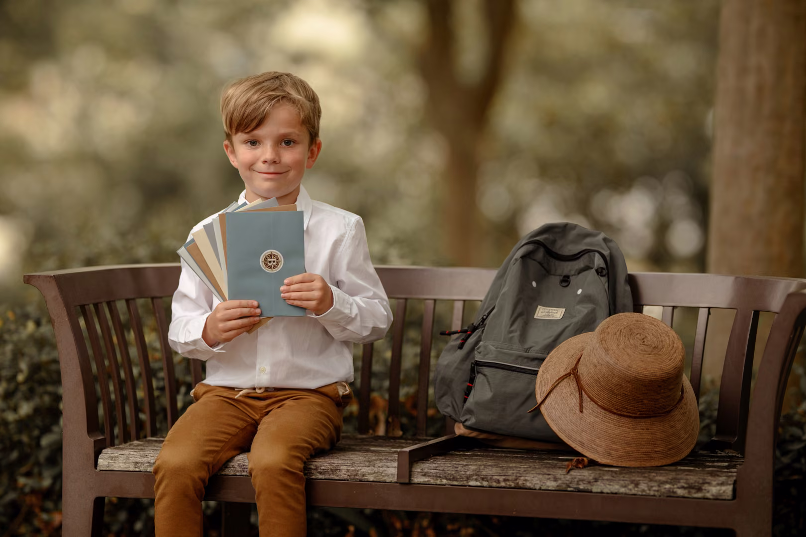 A boy holding small booklets on a wooden bench beside his backpack and hat.